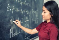 Beautiful Asian student writing on blackboard with chalk in classroom.